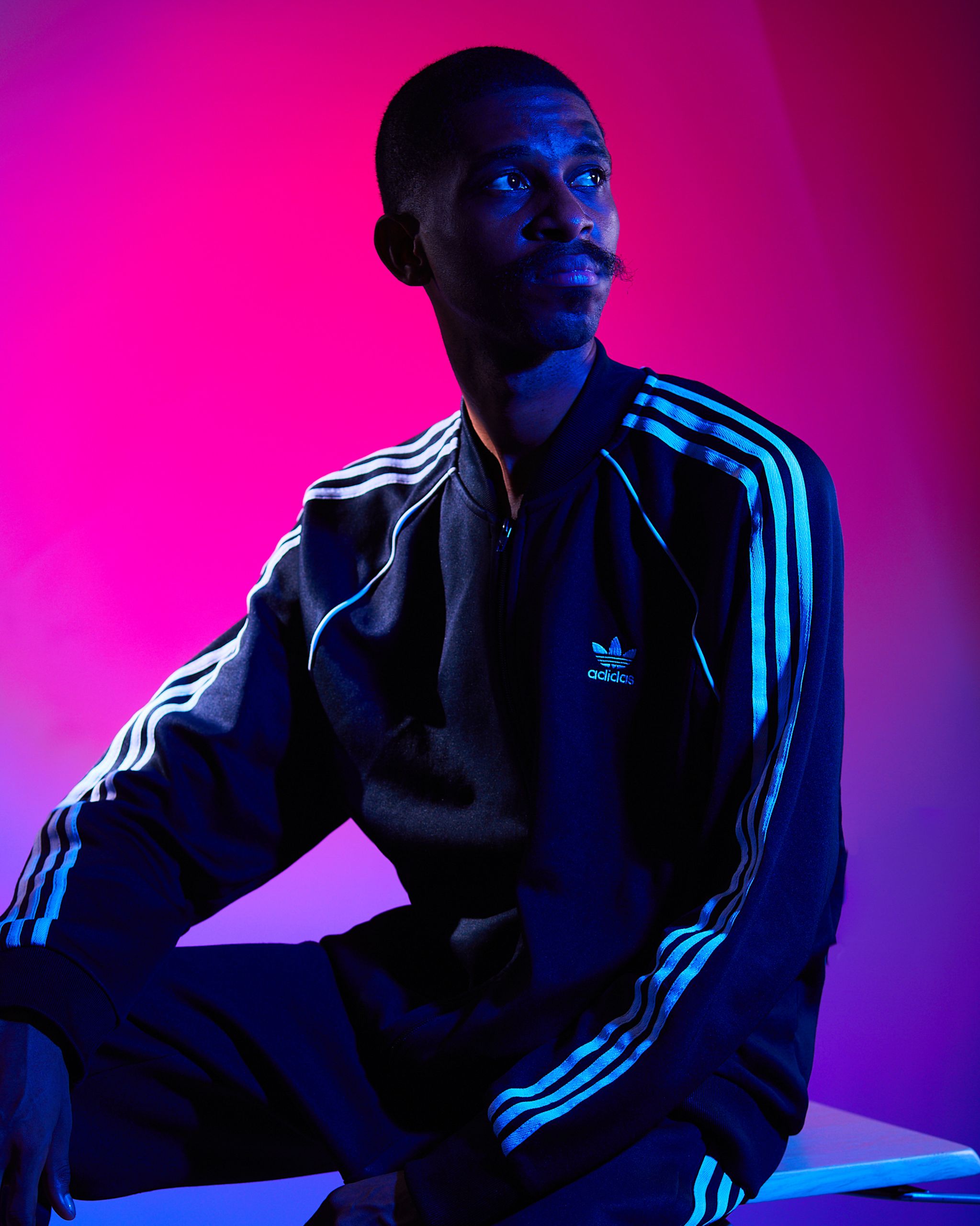 A man in a tracksuit sits under dynamic colored lighting, creating a moody vibe.